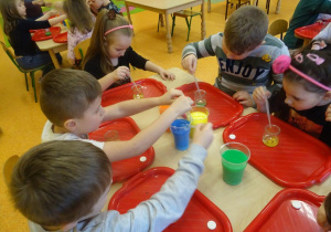 Dzieci nabiera zabarwioną ciesz stojącą w pojemnikach na środku stolika i wlewają ją do słoiczków przed każdym dzieckiem.